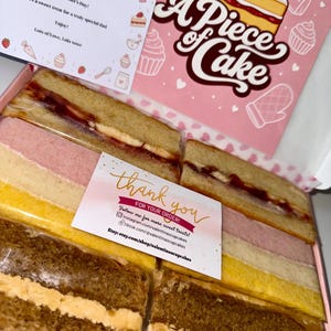 May include: A box of assorted rectangular cake slices featuring strawberry jam, pink, yellow, and brown sponge layers with cream filling. The box lid displays a strawberry cake illustration and the text A Piece of Cake, with two cards included.