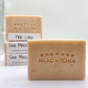 May include: Close-up of several rectangular bars of soap. The soap is a light beige color and has the words "NICKO KITCHEN" and a floral design stamped on the top. Some bars have labels that read "Pink Lotus" and "Sea Moss".