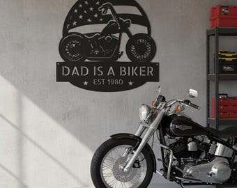 May include: A black metal wall sign featuring a silhouette of a motorcycle overlaid on an American flag design. The text reads DAD IS A BIKER and EST 1980, with a star on each side of the year. The sign is mounted on a light gray wall.