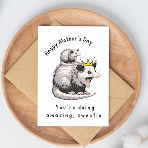 May include: Mother's Day greeting card featuring a cartoon opossum wearing a crown, with a smaller opossum on its head. The card reads "Happy Mother's Day" and "You're doing amazing, sweetie." The card is on a wooden tray with a brown envelope.