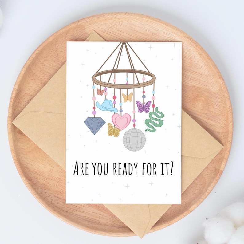 Peut inclure: Carte de v&oelig;ux avec un design fantaisiste repr&eacute;sentant un mobile avec un chapeau de cow-boy, des papillons, un c&oelig;ur, une boule disco et un serpent. Le texte "ARE YOU READY FOR IT?" est imprim&eacute; en dessous. La carte est sur un plateau en bois avec une enveloppe.