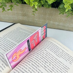 Puede incluir: Un marcap&aacute;ginas rosa y naranja con un dise&ntilde;o de llama y las palabras "Slew Buin" y "Book Club". El marcap&aacute;ginas est&aacute; colocado en un libro con el texto "A Court of Wings and Ruin".