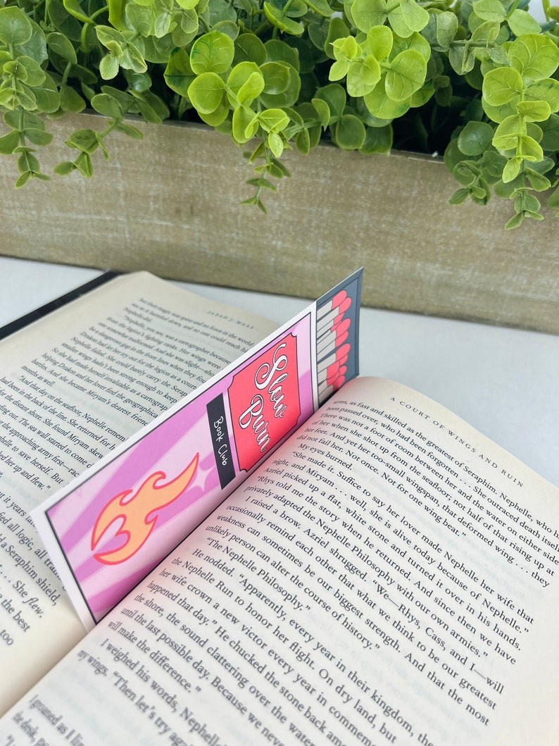 Puede incluir: Un marcap&aacute;ginas rosa y naranja con un dise&ntilde;o de llama y las palabras "Slew Buin" y "Book Club". El marcap&aacute;ginas est&aacute; colocado en un libro con el texto "A Court of Wings and Ruin".