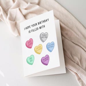 May include: A white card with the text "I hope your birthday is filled with" and six colorful candy hearts with different sayings on them. The sayings are "Spicy Books", "Enemies to Lovers", "Morally Grey Men", "Slow Burn Romance", "One Bed", and "Men Written by Women".