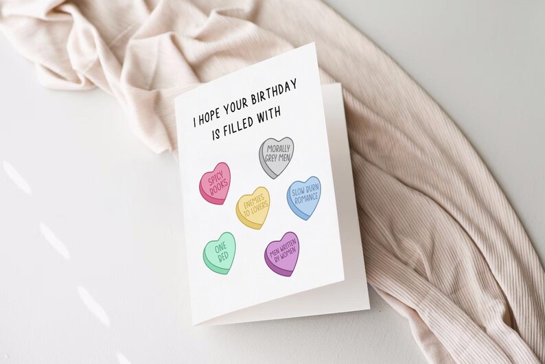 May include: A white card with the text "I hope your birthday is filled with" and six colorful candy hearts with different sayings on them. The sayings are "Spicy Books", "Enemies to Lovers", "Morally Grey Men", "Slow Burn Romance", "One Bed", and "Men Written by Women".