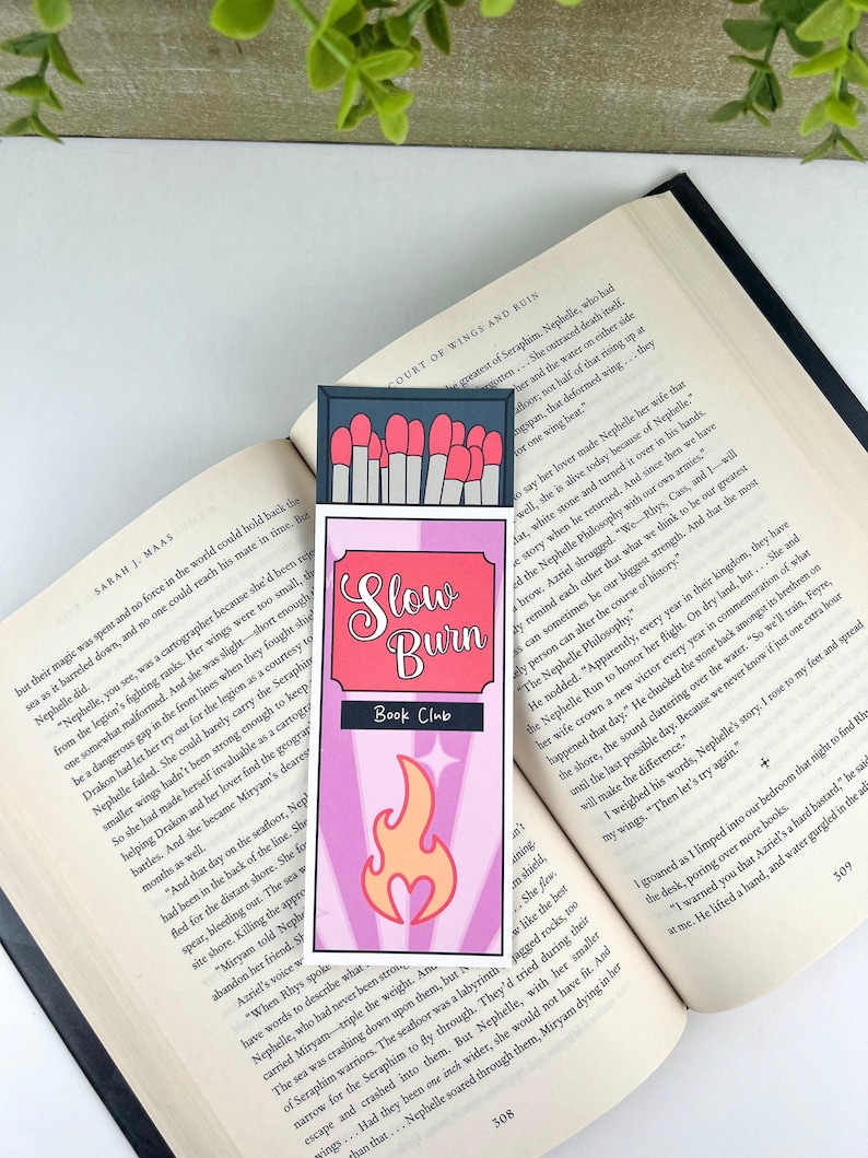 Puede incluir: Un marcap&aacute;ginas rosa y blanco con un dise&ntilde;o de caja de cerillas. La caja de cerillas tiene una llama roja con un coraz&oacute;n en el centro. El texto en el marcap&aacute;ginas dice "Slow Burn Book Club".