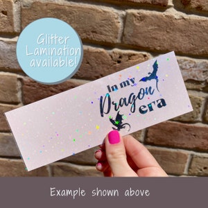 May include: A pink bookmark with iridescent glitter and the text "In my Dragon era" in black.