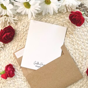 May include: A blank white card with a brown envelope. The card has the text "Golden Hour Graphi" printed on the bottom. The card and envelope are surrounded by white daisies and red carnations.