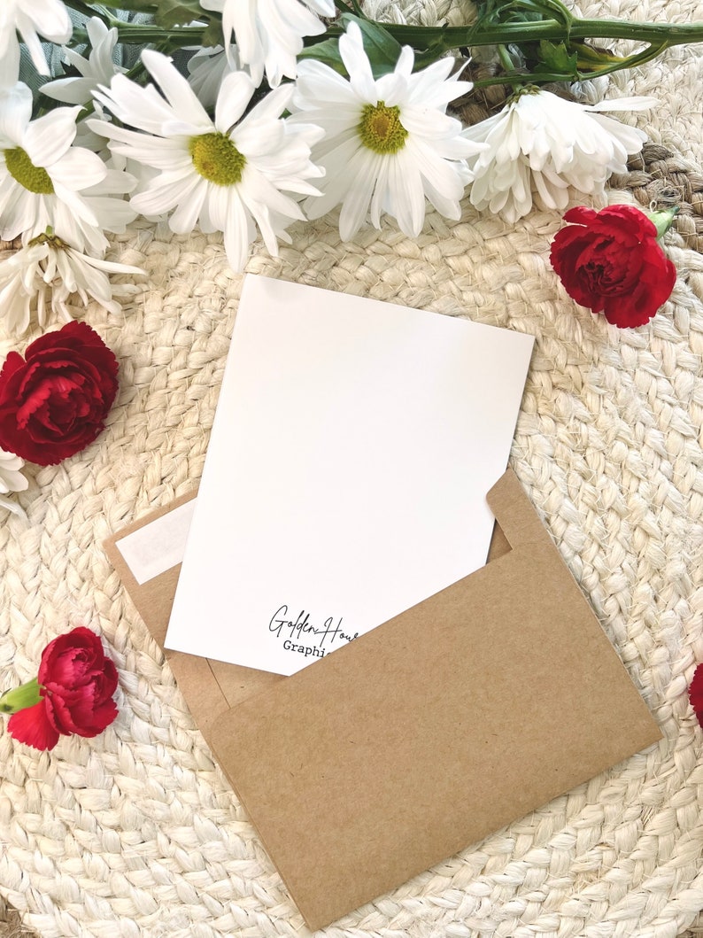 May include: A blank white card with a brown envelope. The card has the text "Golden Hour Graphi" printed on the bottom. The card and envelope are surrounded by white daisies and red carnations.