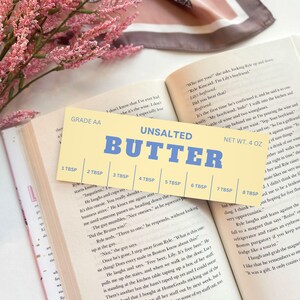 K&ouml;nnte beinhalten: Ein gelbes und blaues Messl&ouml;ffel-Set mit der Aufschrift "Unsalted Butter". Das Messl&ouml;ffel-Set hat Messungen von 1 Essl&ouml;ffel bis 8 Essl&ouml;ffel. Das Messl&ouml;ffel-Set liegt auf einem Buch mit dem Text "Es ist das erste Mal, dass er es best&auml;tigt, und er hat es so selbstbewusst gesagt. "Mein Freund, was?" Ich gehe in die K&uuml;che und nehme eine Flasche Wein und zwei Weingl&auml;ser."