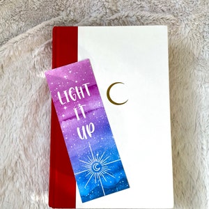 Puede incluir: Un marcap&aacute;ginas de acuarela morado y azul con una luna creciente dorada y estrellas. El marcap&aacute;ginas dice "LIGHT IT UP" en texto blanco.