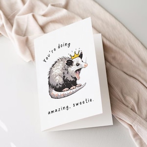 May include: A white card with a black and white illustration of a possum wearing a gold crown. The text on the card reads "You're doing amazing, sweetie."