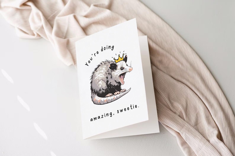 May include: A white card with a black and white illustration of a possum wearing a gold crown. The text on the card reads "You're doing amazing, sweetie."