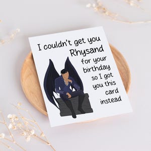 May include: A humorous birthday card featuring a black and white illustration of a character with wings sitting on a throne. The card reads "I couldn't get you Rhysand for your birthday so I got you this card instead."