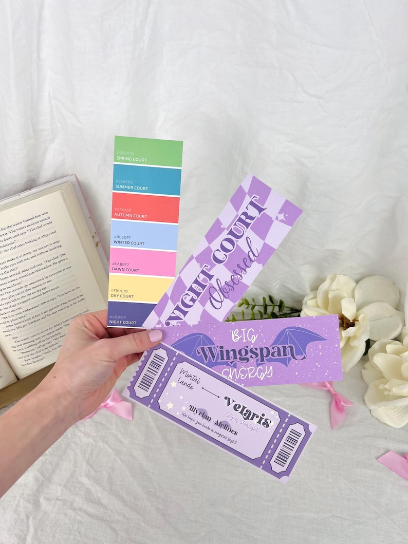 May include: A set of four purple and white bookmarks with a fantasy theme. The bookmarks feature text that reads "Night Court Obsessed", "Big Wingspan Energy", and "Velaris Illyrian Airlines".
