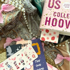 Puede incluir: Primer plano de un libro titulado "It Starts With Us" de Colleen Hoover, con una portada rosa y dorada. El libro est&aacute; rodeado de varios marcadores con diferentes dise&ntilde;os, incluyendo un marcador rosa con fresas, un marcador azul con un marco de fotos y un marcador blanco y negro con una mariposa.