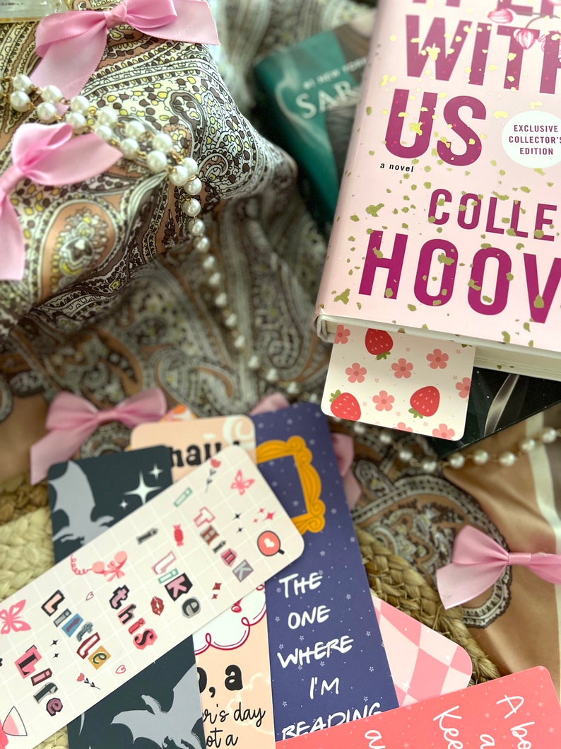 Puede incluir: Primer plano de un libro titulado "It Starts With Us" de Colleen Hoover, con una portada rosa y dorada. El libro est&aacute; rodeado de varios marcadores con diferentes dise&ntilde;os, incluyendo un marcador rosa con fresas, un marcador azul con un marco de fotos y un marcador blanco y negro con una mariposa.