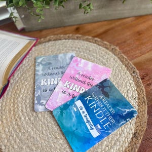 May include: Three rectangular bookmarks with the text "A reader without their Kindle is a tragedy." The bookmarks have a marble design in shades of gray, pink, and blue. They are on a woven, circular placemat.