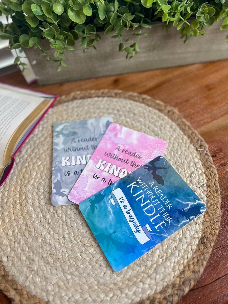May include: Three rectangular bookmarks with the text "A reader without their Kindle is a tragedy." The bookmarks have a marble design in shades of gray, pink, and blue. They are on a woven, circular placemat.