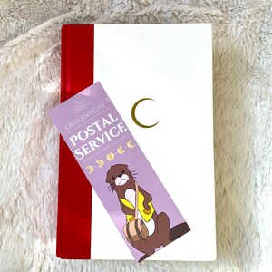 Puede incluir: Un diario blanco con una encuadernaci&oacute;n roja y un marcap&aacute;ginas. El marcap&aacute;ginas es morado con una nutria de dibujos animados que lleva una bolsa y el texto "Crescent City Postal Service".