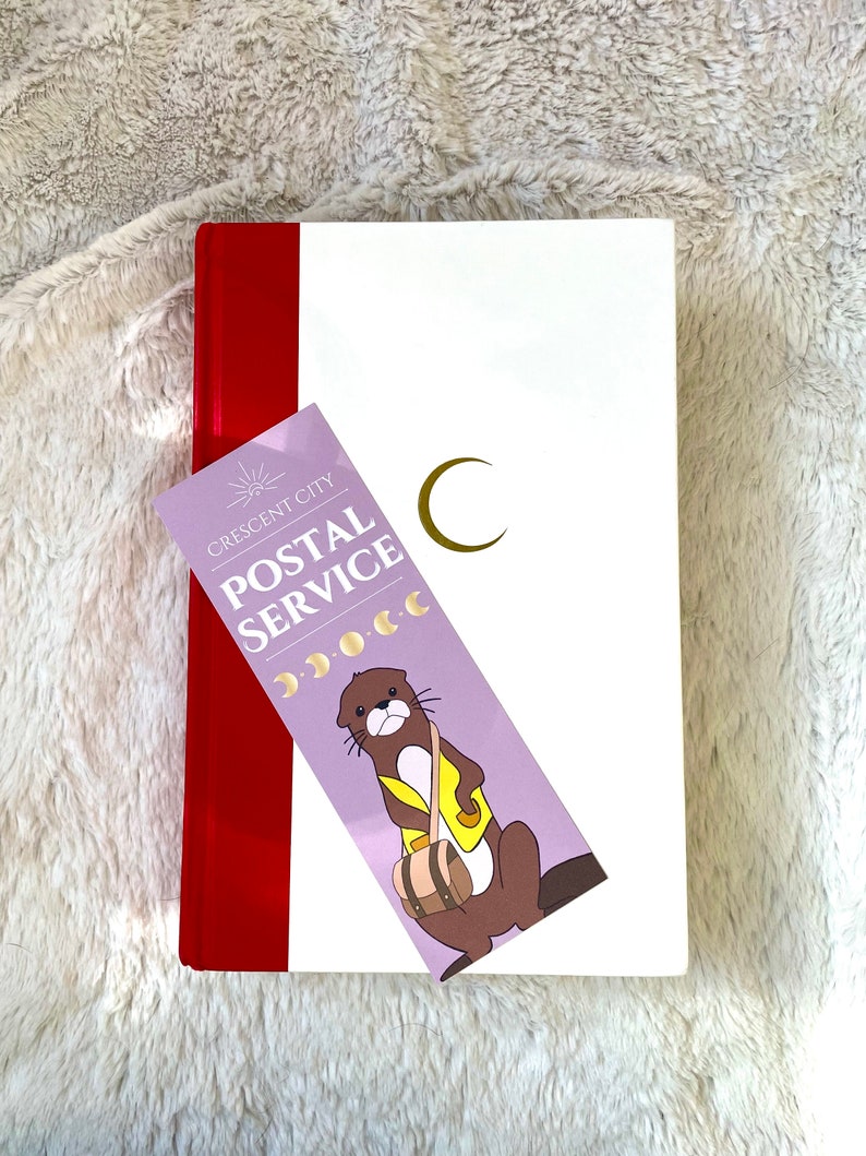 Puede incluir: Un diario blanco con una encuadernaci&oacute;n roja y un marcap&aacute;ginas. El marcap&aacute;ginas es morado con una nutria de dibujos animados que lleva una bolsa y el texto "Crescent City Postal Service".