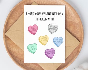 May include: A Valentine's Day greeting card with the message "I hope your Valentine's Day is filled with" and candy heart graphics. The hearts have phrases like "Spicy Books" and "Slow Burn Romance" on them. The card is on a wooden tray.