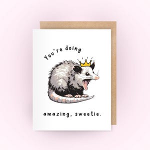 May include: A greeting card with a cartoon possum wearing a gold crown. The text on the card reads "You're doing amazing, sweetie."