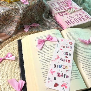 Puede incluir: Un marcap&aacute;ginas rosa y blanco con el texto "I think I like this little life" y varias ilustraciones, incluyendo una mariposa, una fresa, una copa de vino y corazones. El marcap&aacute;ginas est&aacute; encima de un libro abierto con una cinta rosa.