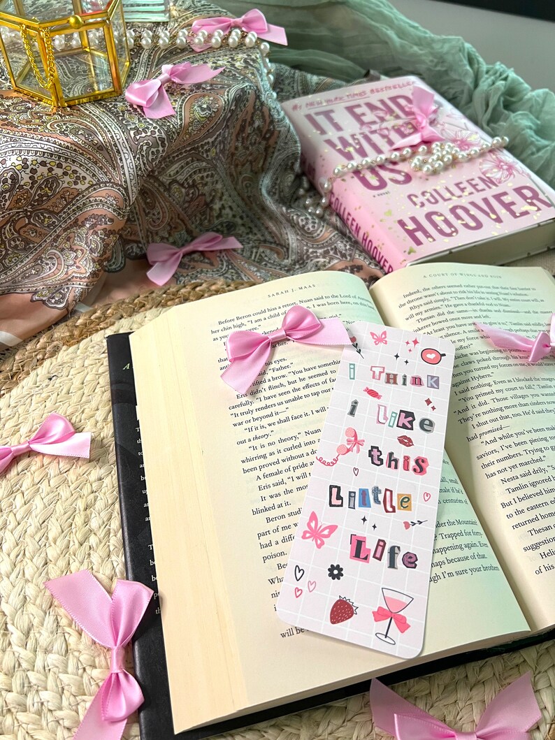 Puede incluir: Un marcap&aacute;ginas rosa y blanco con el texto "I think I like this little life" y varias ilustraciones, incluyendo una mariposa, una fresa, una copa de vino y corazones. El marcap&aacute;ginas est&aacute; encima de un libro abierto con una cinta rosa.