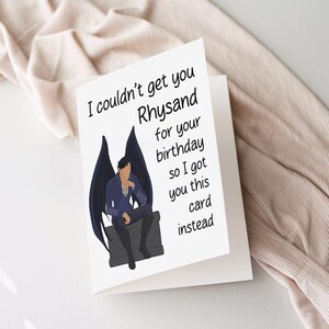 May include: A white card with black text that reads "I couldn't get you Rhysand for your birthday so I got you this card instead."  The card features a black and white illustration of a man with wings sitting on a stone.