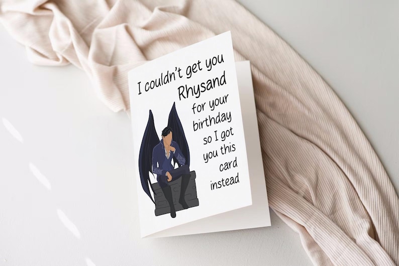 May include: A white card with black text that reads "I couldn't get you Rhysand for your birthday so I got you this card instead."  The card features a black and white illustration of a man with wings sitting on a stone.