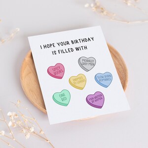 May include: A birthday card with the text "I hope your birthday is filled with" and a series of candy hearts with different sayings, including "Spicy Books", "One Red", "Enemies to Lovers", "Morally Grey Men", "Men Written by Women", and "Slow Burn Romance".