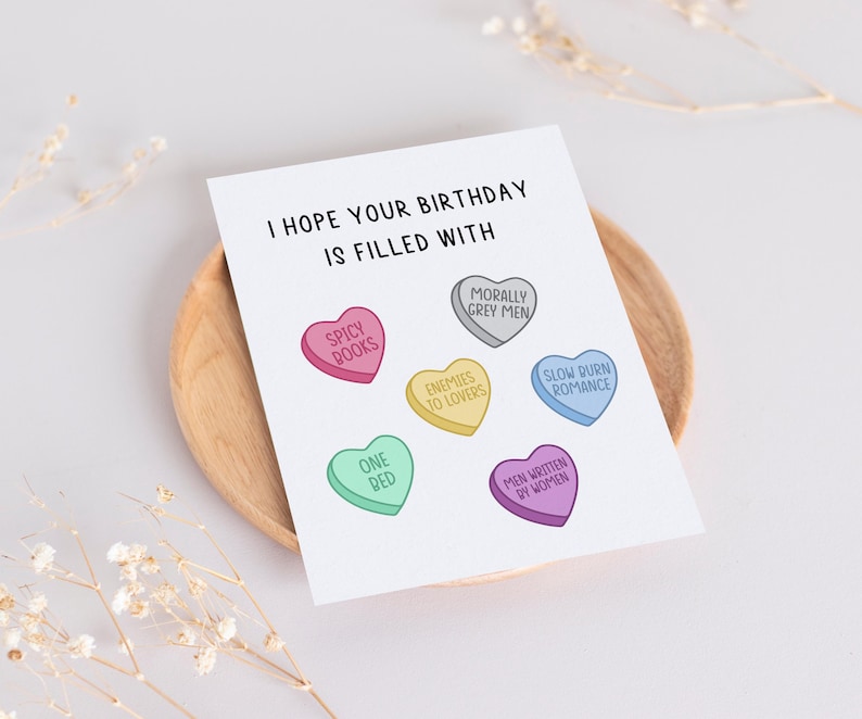 May include: A birthday card with the text "I hope your birthday is filled with" and a series of candy hearts with different sayings, including "Spicy Books", "One Red", "Enemies to Lovers", "Morally Grey Men", "Men Written by Women", and "Slow Burn Romance".