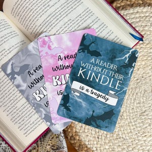 May include: A stack of four rectangular bookmarks with the text "A reader without their Kindle is a tragedy." The bookmarks have different backgrounds: gray, pink, and blue with dragon silhouettes.