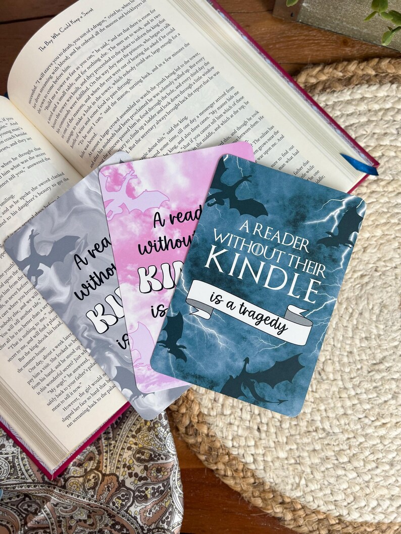 May include: A stack of four rectangular bookmarks with the text "A reader without their Kindle is a tragedy." The bookmarks have different backgrounds: gray, pink, and blue with dragon silhouettes.