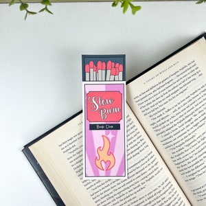 Puede incluir: Un marcap&aacute;ginas rosa y morado con un dise&ntilde;o de caja de cerillas. El marcap&aacute;ginas presenta una llama con un coraz&oacute;n en el centro y el texto "Slow Burn Book Club".
