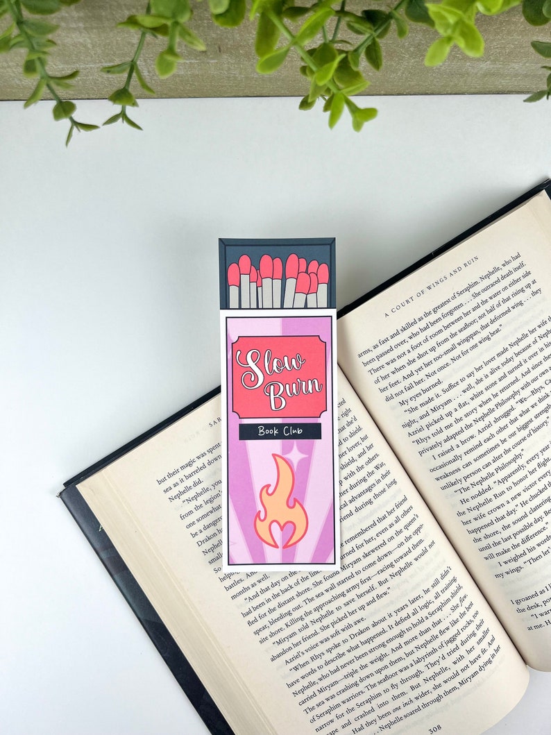 Puede incluir: Un marcap&aacute;ginas rosa y morado con un dise&ntilde;o de caja de cerillas. El marcap&aacute;ginas presenta una llama con un coraz&oacute;n en el centro y el texto "Slow Burn Book Club".