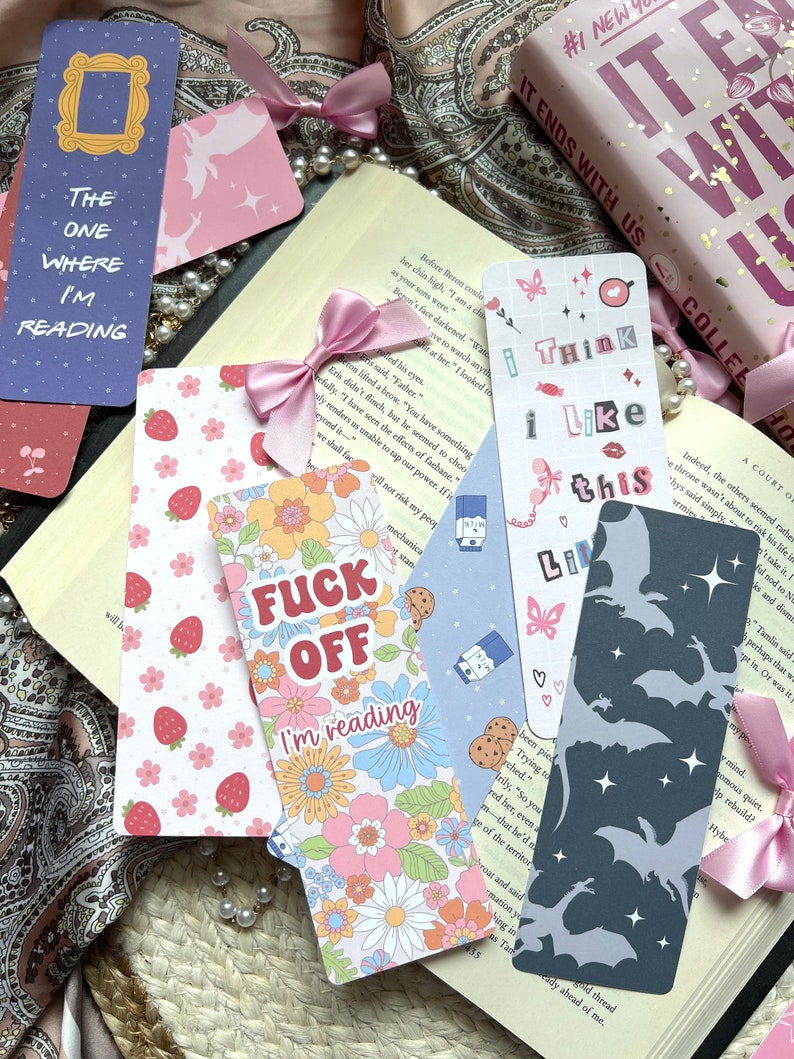 May include: A collection of colorful bookmarks with various designs. One bookmark features a floral pattern with the text "I'm reading". Another bookmark has a pink and white strawberry pattern. A third bookmark has a blue and white pattern with the text "Think I Like This". A fourth bookmark has a gray silhouette of bats with stars. A fifth bookmark has a blue and white pattern with the text "The One Where I'm Reading".