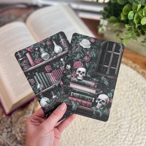 May include: Two rectangular cards with gothic illustrations. One card depicts a bookshelf with potion bottles and roses. The other card shows a skull, books, and a window. The cards are held in a hand, with a book and greenery in the background.
