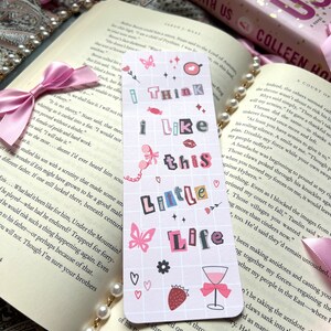 Puede incluir: Un marcap&aacute;ginas rosa y blanco con un patr&oacute;n de cuadr&iacute;cula e ilustraciones coloridas. El marcap&aacute;ginas presenta una mariposa, una fresa, una copa de vino y corazones. El texto en el marcap&aacute;ginas dice "I think I like this little life".