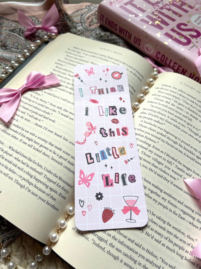 Puede incluir: Un marcap&aacute;ginas rosa y blanco con un patr&oacute;n de cuadr&iacute;cula e ilustraciones coloridas. El marcap&aacute;ginas presenta una mariposa, una fresa, una copa de vino y corazones. El texto en el marcap&aacute;ginas dice "I think I like this little life".