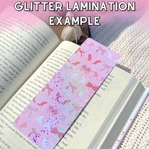 K&ouml;nnte beinhalten: Ein rosa-wei&szlig;es Lesezeichen mit Glitzer-Finish. Das Lesezeichen zeigt ein sich wiederholendes Muster aus Flederm&auml;usen und Schleifen. Der Text "GLITTER LAMINATION EXAMPLE" ist oben im Bild gedruckt.