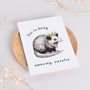 May include: A greeting card with a cartoon possum wearing a crown. The text on the card reads "You're doing amazing, sweetie."