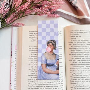 May include: A book bookmark featuring a portrait of a woman blowing a bubble. The bookmark is printed on a light purple background with a checkered pattern. The text "You're really ugh! This day" is visible on the page behind the bookmark.