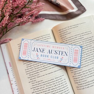 May include: A close-up of a blue and white ticket-shaped bookmark with the text "Lifetime Member Jane Austen Book Club". The bookmark is lying on top of an open book with text about a boyfriend.