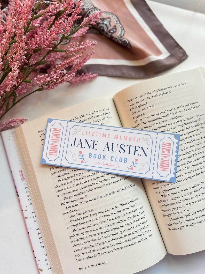 May include: A close-up of a blue and white ticket-shaped bookmark with the text "Lifetime Member Jane Austen Book Club". The bookmark is lying on top of an open book with text about a boyfriend.