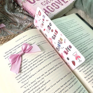 Puede incluir: Un marcap&aacute;ginas rosa y blanco con el texto "Think Like This" y varias pegatinas de colores. El marcap&aacute;ginas est&aacute; decorado con una cinta rosa y se coloca encima de un libro titulado "The Witch of Us" de Colleen Hoover.