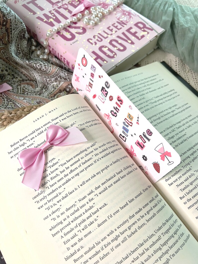 Puede incluir: Un marcap&aacute;ginas rosa y blanco con el texto "Think Like This" y varias pegatinas de colores. El marcap&aacute;ginas est&aacute; decorado con una cinta rosa y se coloca encima de un libro titulado "The Witch of Us" de Colleen Hoover.