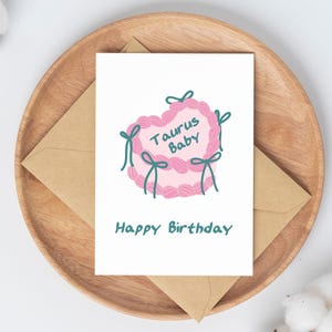 May include: A white card with a pink heart-shaped cake with green frosting and the text "Taurus Baby" on it. The card also has the text "Happy Birthday" in green.