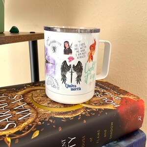 Puede incluir: Taza de viaje blanca con un gr&aacute;fico en blanco y negro de alas y una espada, con el texto "Umbra mortis" y "Light it up". La taza tambi&eacute;n tiene otros gr&aacute;ficos, incluyendo un personaje con una espada, un personaje con una corona y un personaje con una llama. La taza est&aacute; encima de una pila de libros con el t&iacute;tulo "Crescent City" y "House of Flame and Shadow".